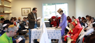 A daytime megillah reading in a living room, with men and women separated by a chest high mechitza and a table. At the table in the center a woman reads the megillah, with a male and female gabbai on either side, following along. Some adults and children are in costume. (frontview)